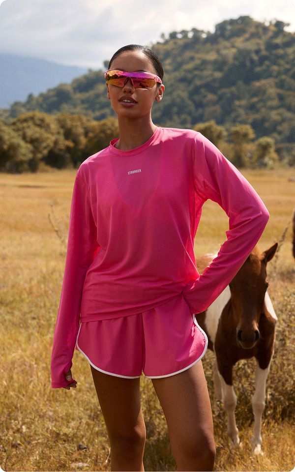 A person in a bright pink athletic outfit and mirrored sunglasses stands in a grassy field with a horse in the background.