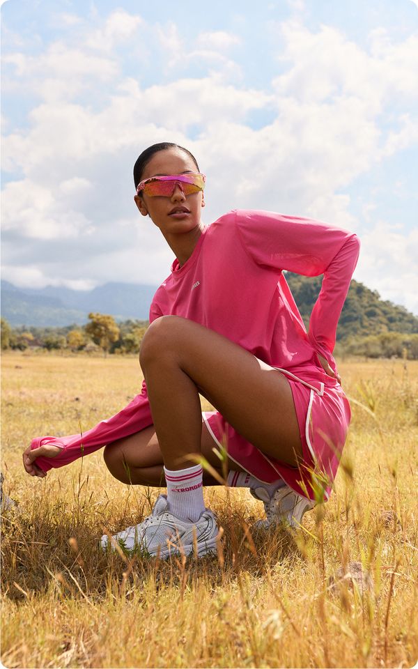 A person in a bright pink athletic outfit and reflective sunglasses squats in a grassy field, with mountains and a cloudy sky in the background.
