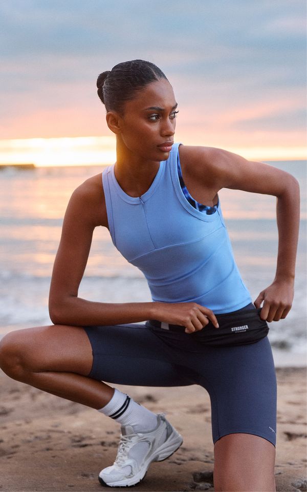 Athletic woman in blue workout gear and white sneakers, kneeling on the beach at sunset, with a focused expression.