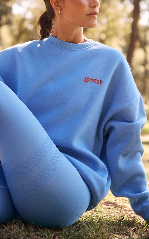 Woman in blue sweatshirt with "STRONGER" logo and matching leggings, sitting outdoors on grass, with blurred trees in background.