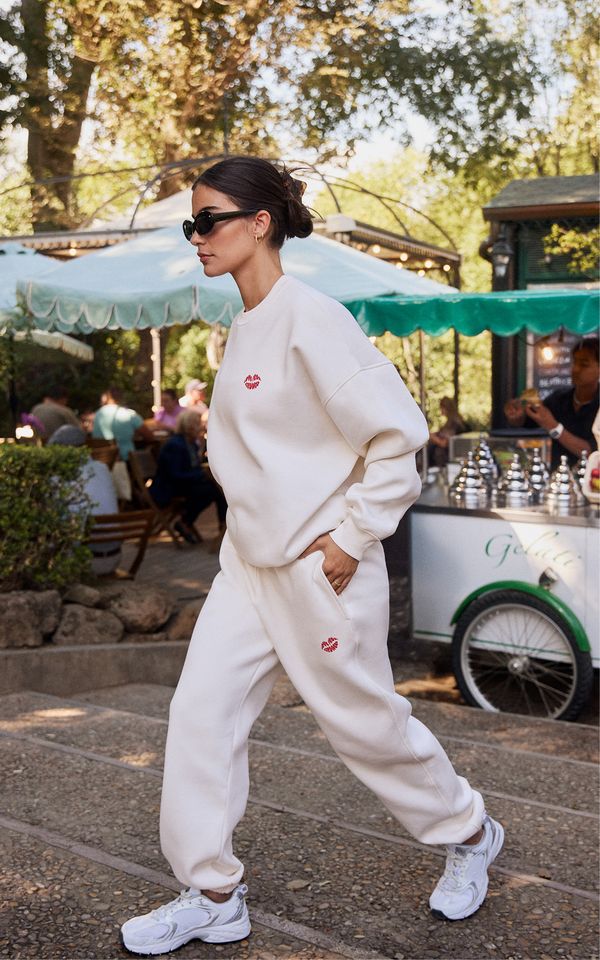 Woman in a cream tracksuit and sunglasses walks outdoors, with a food cart and seated people in the background.