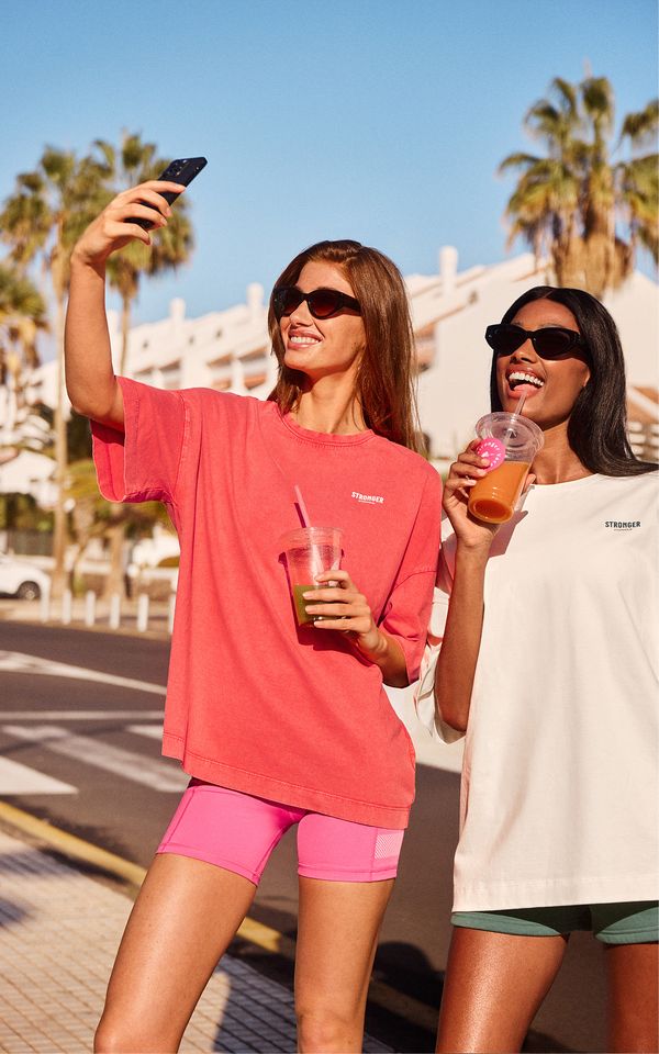Two people in sunglasses taking a selfie and enjoying drinks on a sunny street, with palm trees and buildings in the background.