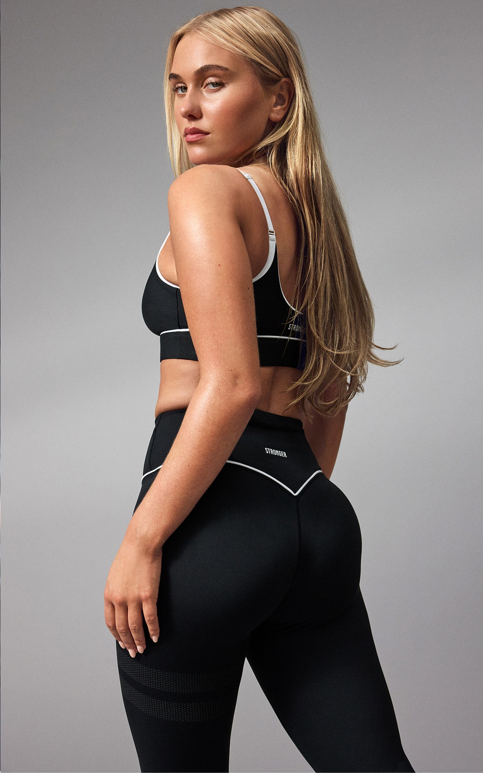 A woman with long blonde hair wearing a black workout outfit poses confidently, turned slightly to the side against a gray background.