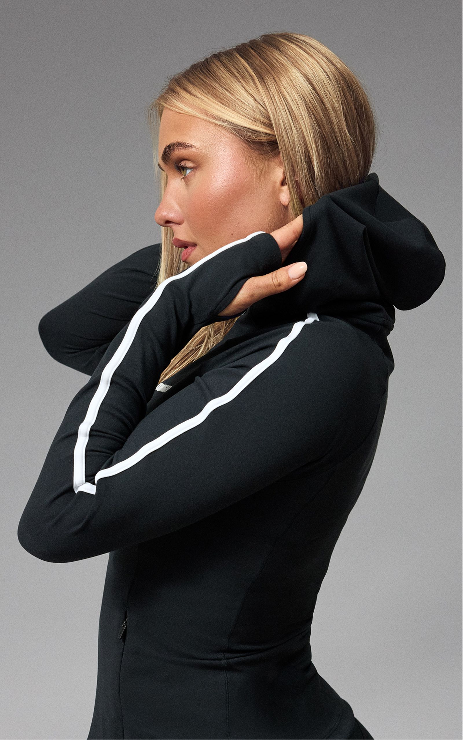 A person wearing a black hoodie with white stripes adjusts the hood. The background is neutral gray.