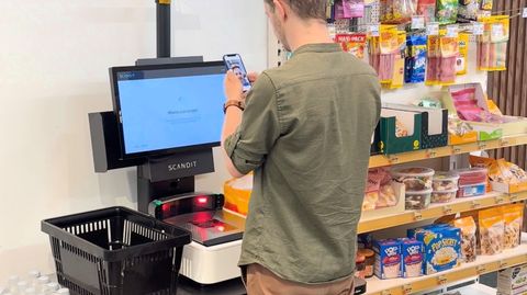 Man using smartphone at a Scandit self-checkout kiosk in a grocery store, with shelves of snacks and a shopping basket nearby.