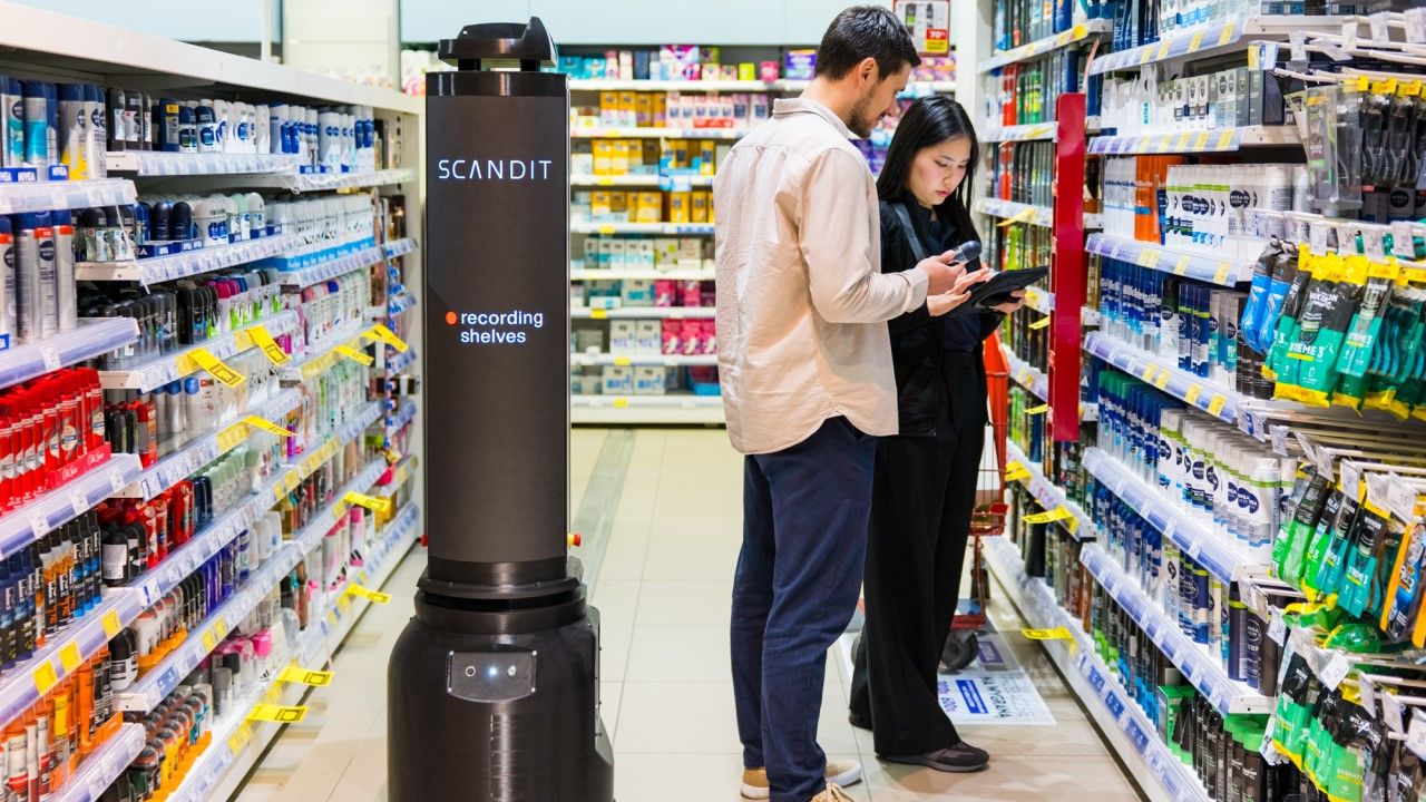 A Scandit inventory robot scans product shelves in a store aisle while a man and woman shop nearby, looking at a device.