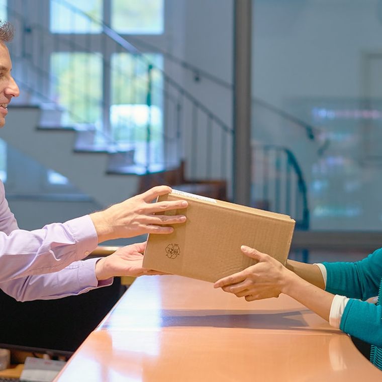 A man in a light shirt hands a package to a smiling woman in a teal cardigan over a counter in an office setting.