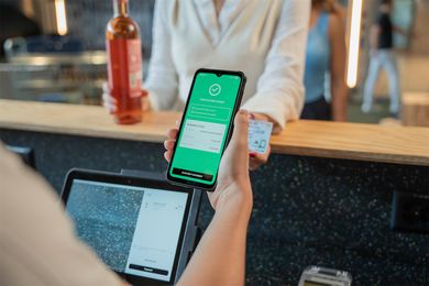 Cashier holding a smartphone displaying a green age and identity verification screen at a bar counter, with a customer holding an ID card in the background.