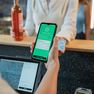 Cashier holding a smartphone displaying a green age and identity verification screen at a bar counter, with a customer holding an ID card in the background.
