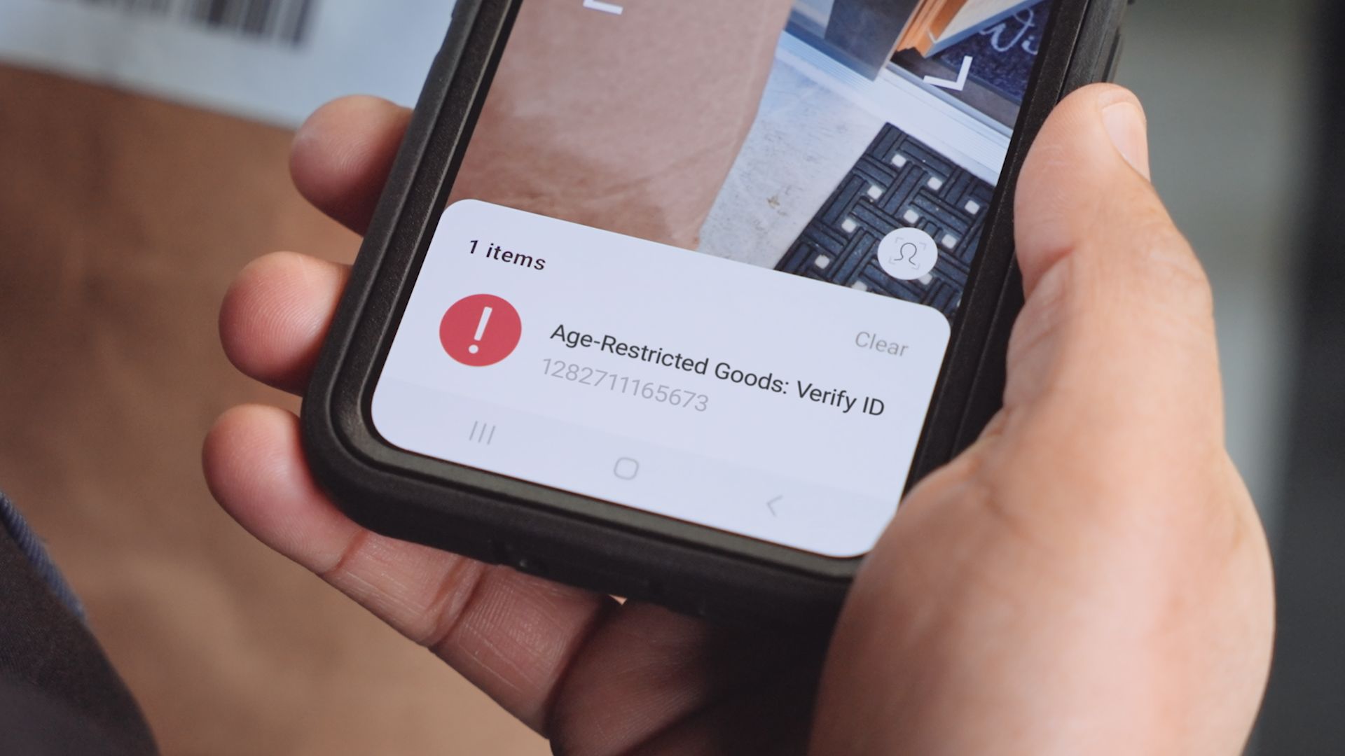 A person holds a phone displaying a notification: "Age-Restricted Goods: Verify ID" with a red exclamation mark icon.