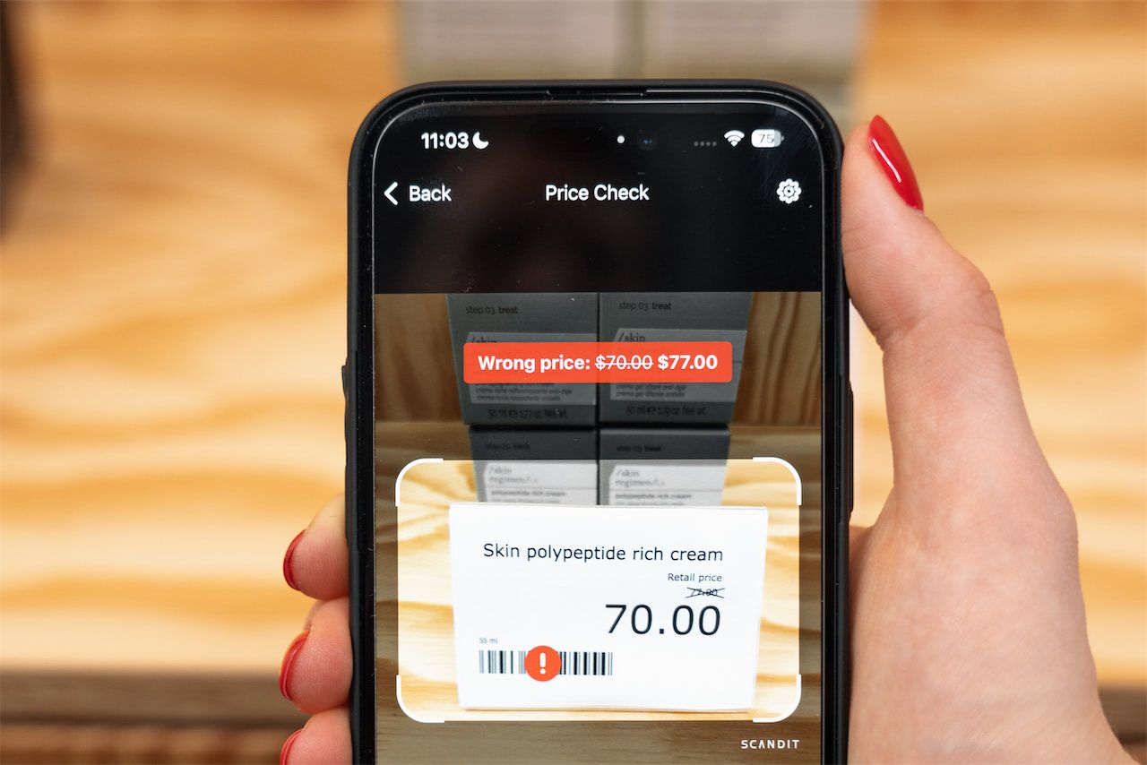 A person holds a smartphone using a price check app. The screen displays an augmented reality view with a "Wrong price" error on a skin cream product.