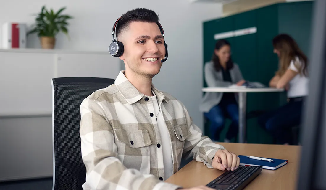 Man wearing headphones with a microphone sits at a desk, smiling at a computer screen. Two people converse in the background.