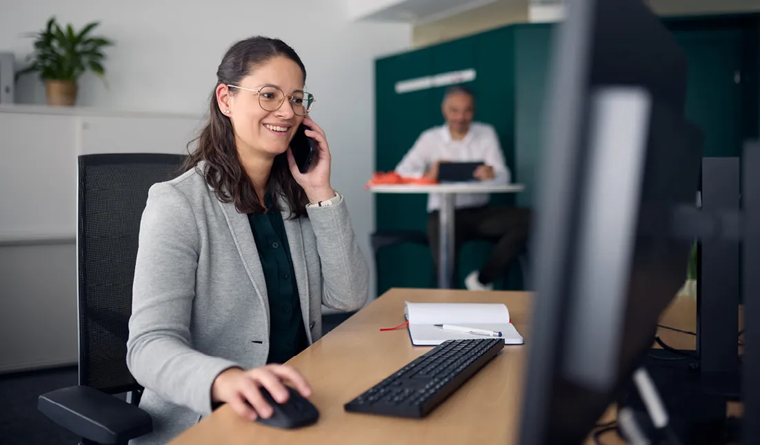 A woman in an office talks on the phone while using a computer. A man works in the background at a table.