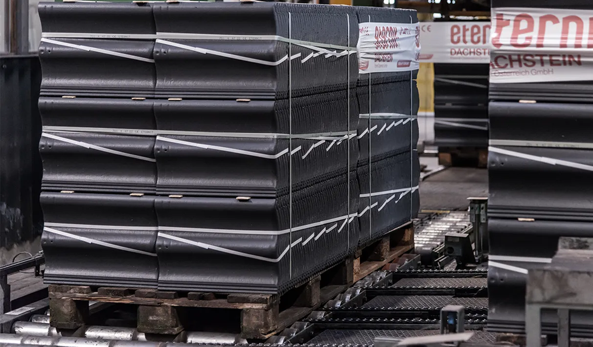 Stacked black roof tiles, neatly arranged and strapped on a wooden pallet; pallet placed on a conveyor system in an industrial setting.