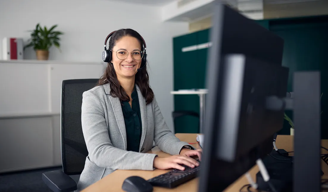 Smiling person with headphones working at a computer in an office setting.