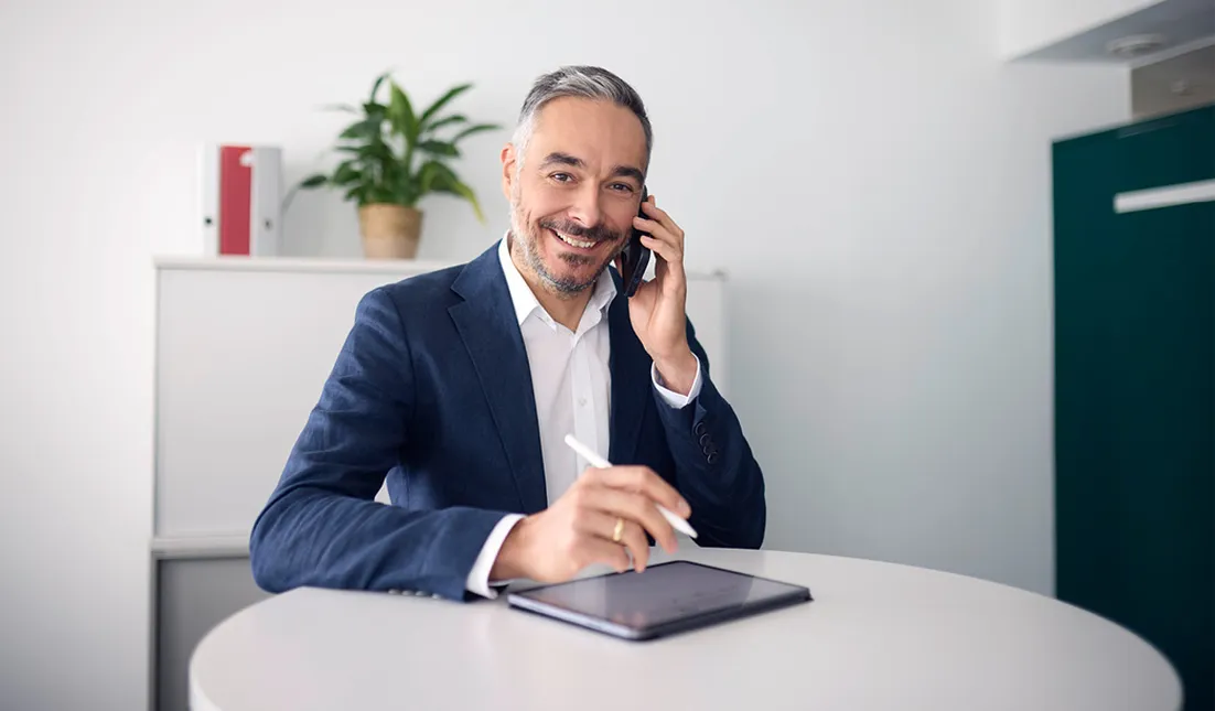 Man in a suit smiling, talking on a phone, and using a tablet with a stylus at a round table in a modern office.