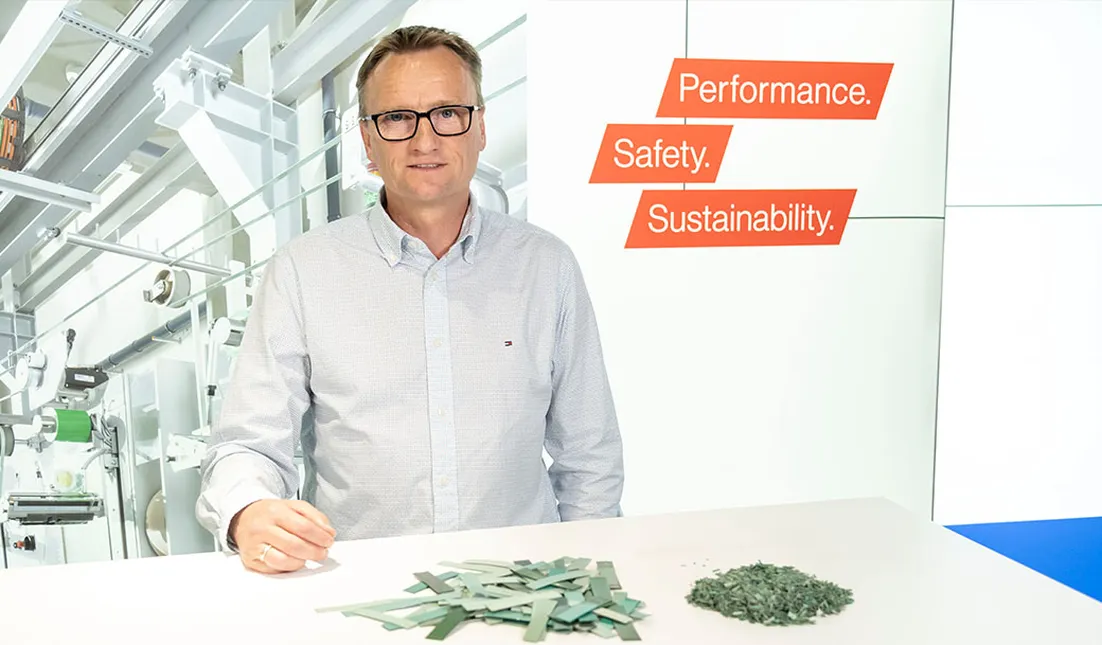Person in a light blue shirt in front of an industrial background, presenting two piles of green material – long strips and shredded pieces – with three orange text boxes reading “Performance.”, “Safety.”, “Sustainability.”.