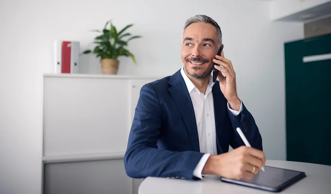 Smiling man in a suit talks on the phone while taking notes at a desk, with a plant and office cabinet in the background.