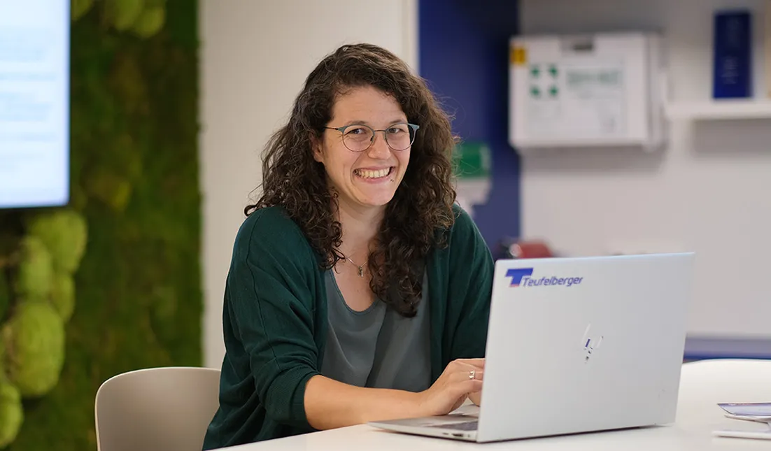 A person with curly hair and glasses smiles while working on a laptop in a modern office setting.