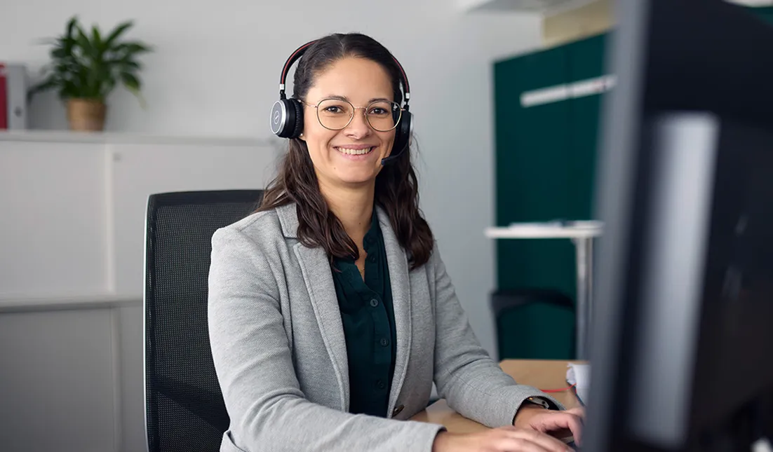 Smiling woman in a blue blazer and headphones sits at a desk in an office, ready to assist customers.