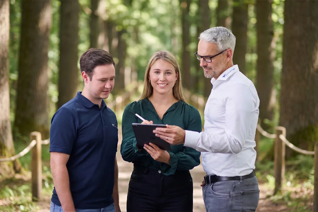 Three people standing in a forest, discussing something on a tablet. The setting is serene, with trees lining the path.