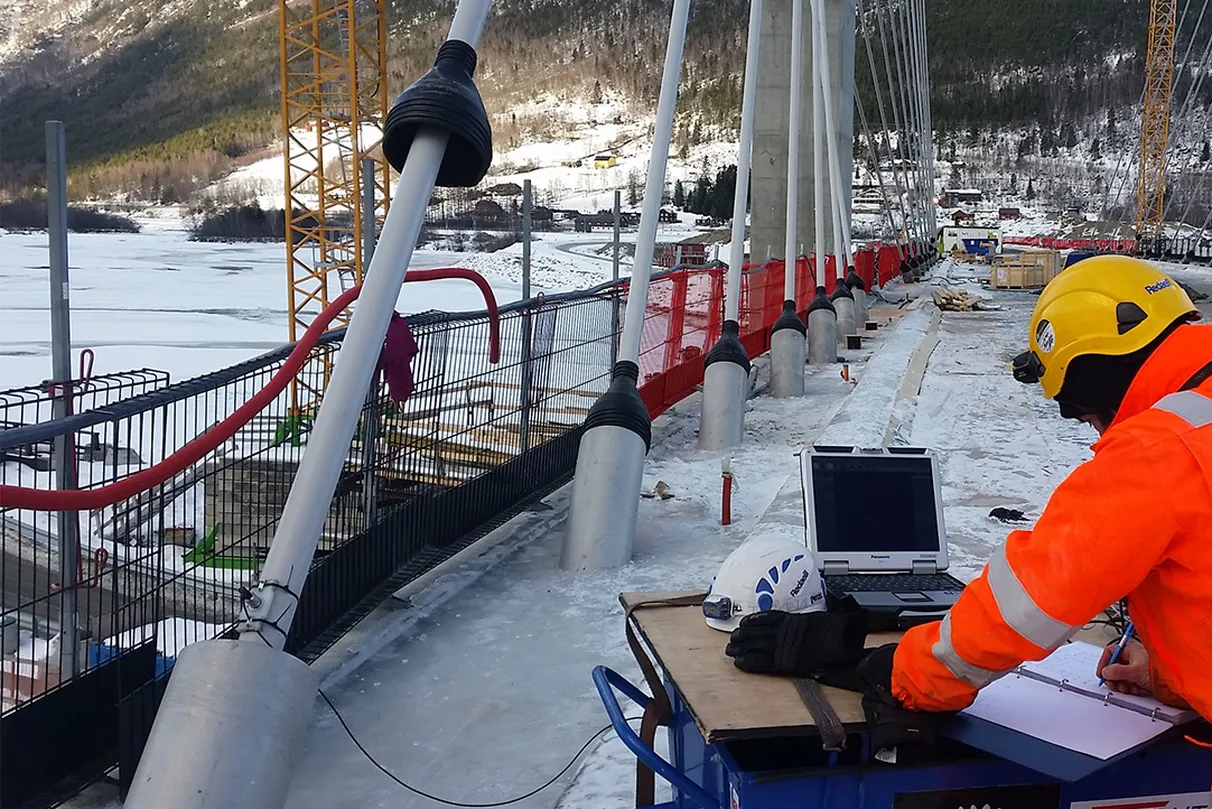 Worker in orange gear and helmet uses a laptop on a snowy bridge under construction, with cranes and mountains in the background.
