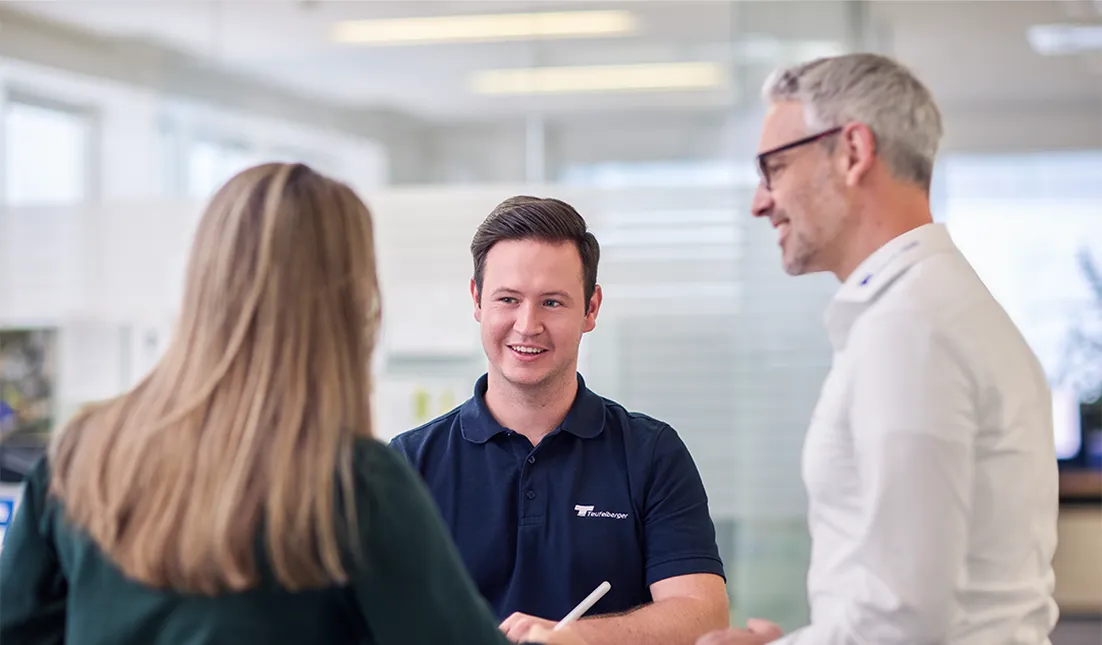 Three people in a modern office having a friendly discussion around a table, with one person writing on a notepad.