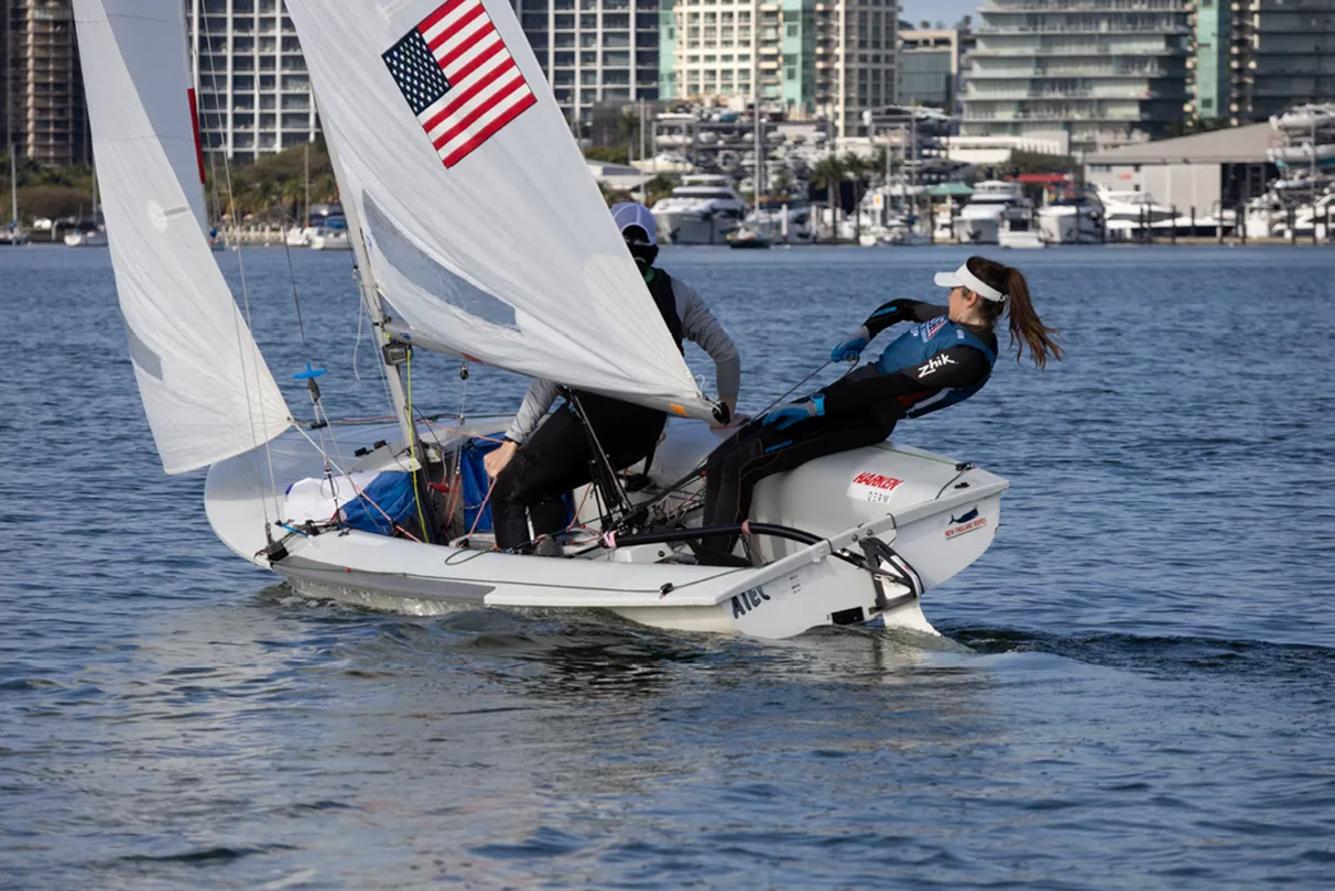 Two people sailing a small boat on calm water, with city buildings in the background. An American flag is visible on the sail.