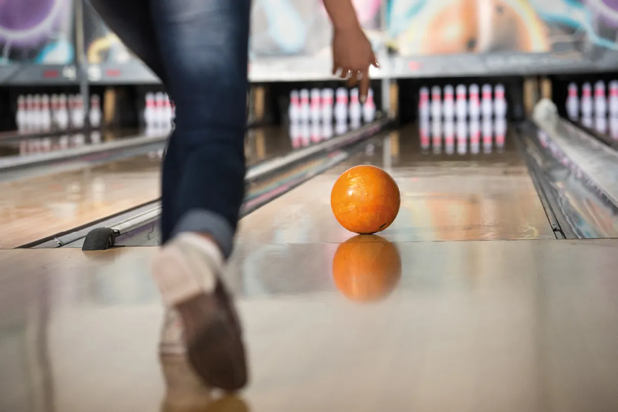 A person in jeans bowls a bright orange ball down a lane towards a set of pins.