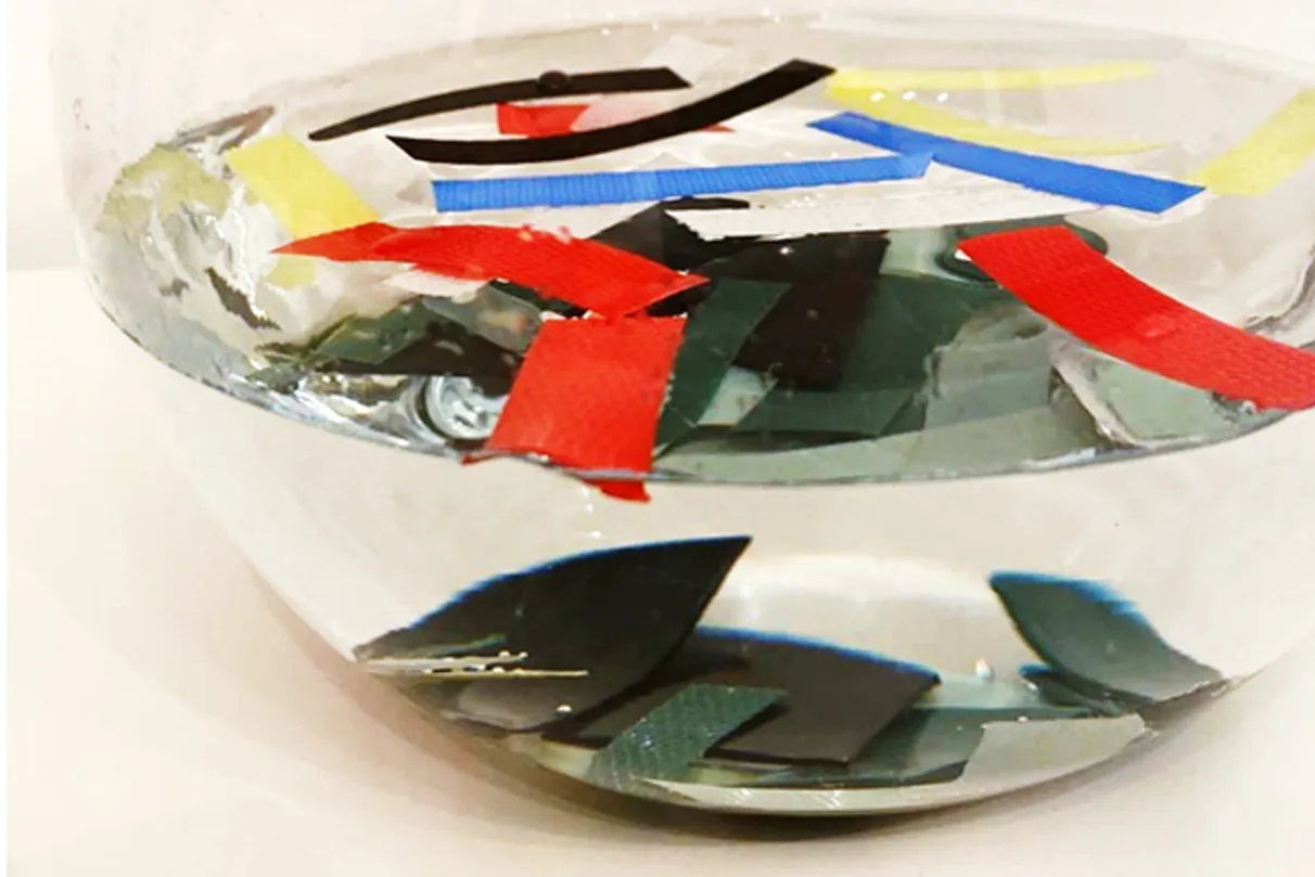 Glass bowl filled with water containing floating and submerged strips of colored strapping – in red, yellow, blue, black, and white.