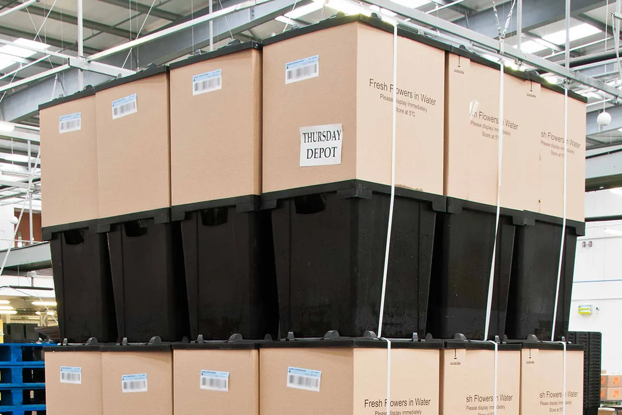 Stack of large cardboard boxes on black plastic pallets in a warehouse; labels read “Fresh Flowers in Water – Please display immediately – Store at 5 °C” and “THURSDAY DEPOT”.