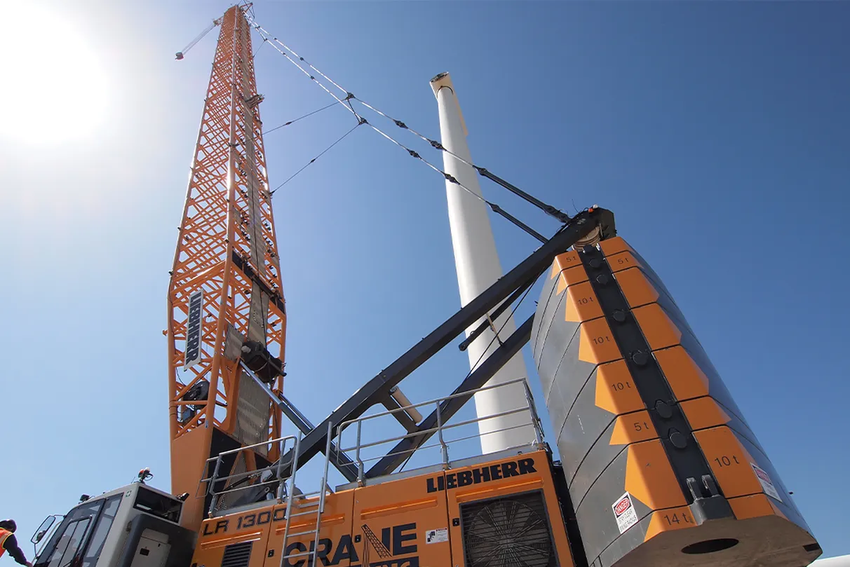 A large orange crane with "Liebherr" branding is lifting a tall white wind turbine part against a clear blue sky.