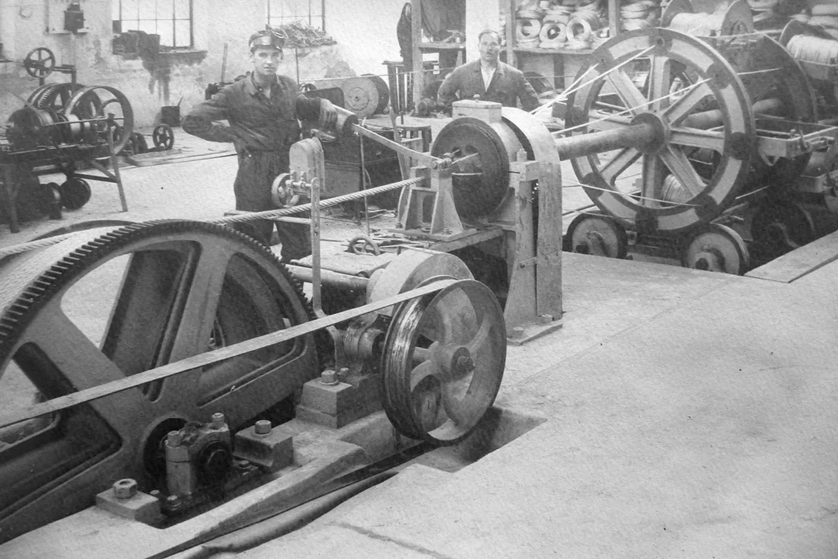 Historical industrial scene showing the production of a fiber rope, with two individuals operating large mechanical equipment featuring gears, belts, and wheels.