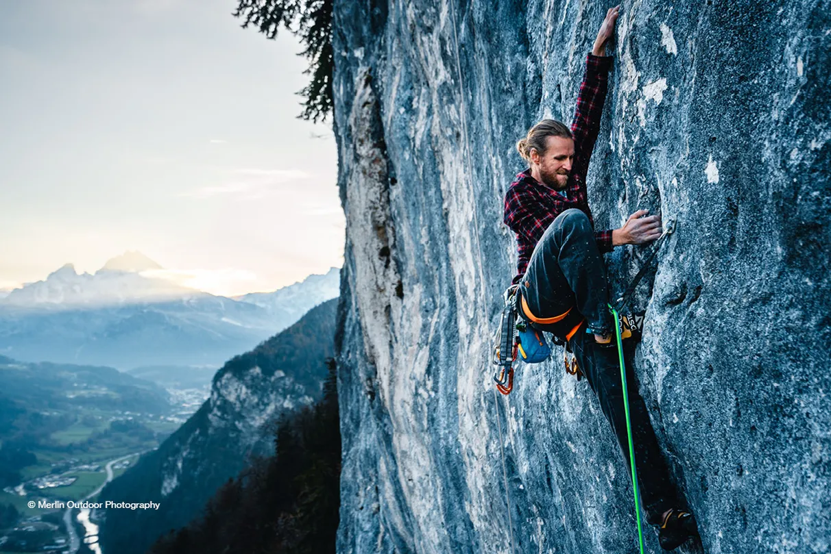 A climber ascends a steep rock face with safety gear, overlooking a scenic valley and mountains.