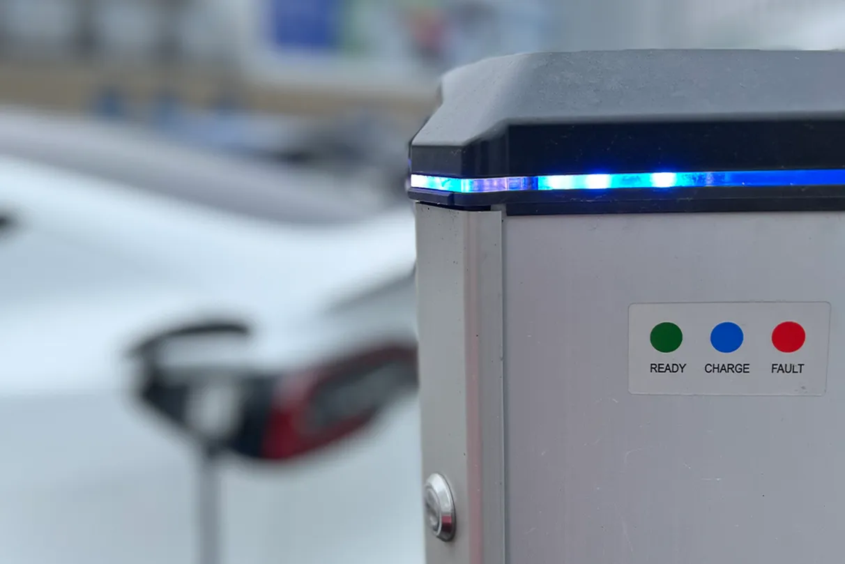 Close-up of an electric vehicle charging station with a blue LED light strip and status indicators. A white EV is connected in the blurred background.