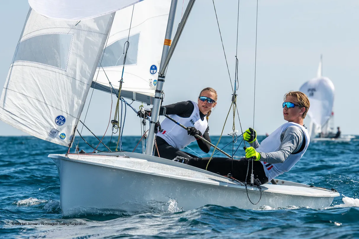 Two sailors maneuver a small sailboat on open water, with sails catching the wind. Another sailboat is visible in the background.
