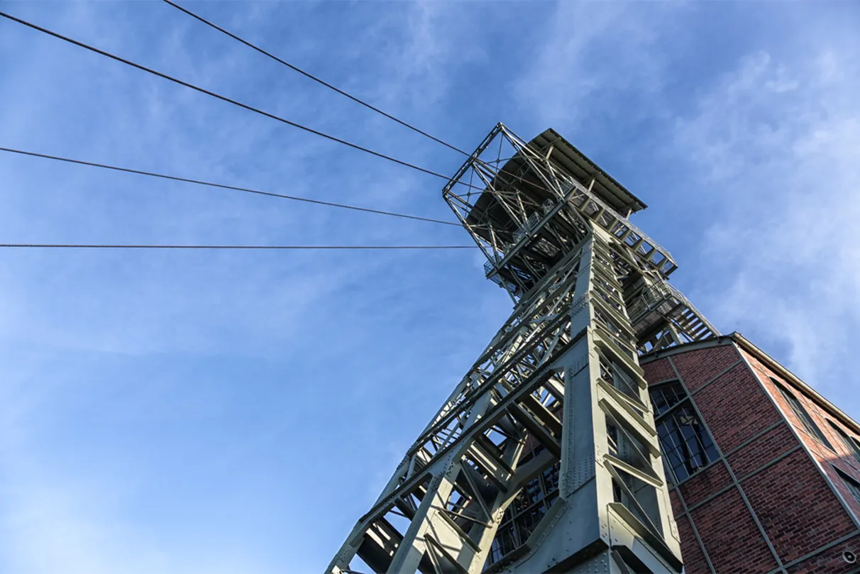 A tall metal industrial structure, likely a mining headframe or hoist tower, with several cables attached, set against a partly cloudy blue sky. A brick building is visible on the right.