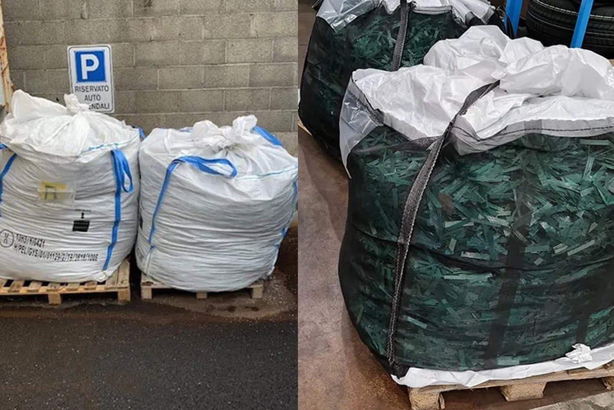 Two large white bags with blue straps on pallets next to two black mesh bags filled with shredded green material – a sign in the background reads “RISERVATO AUTO ANDALU”.