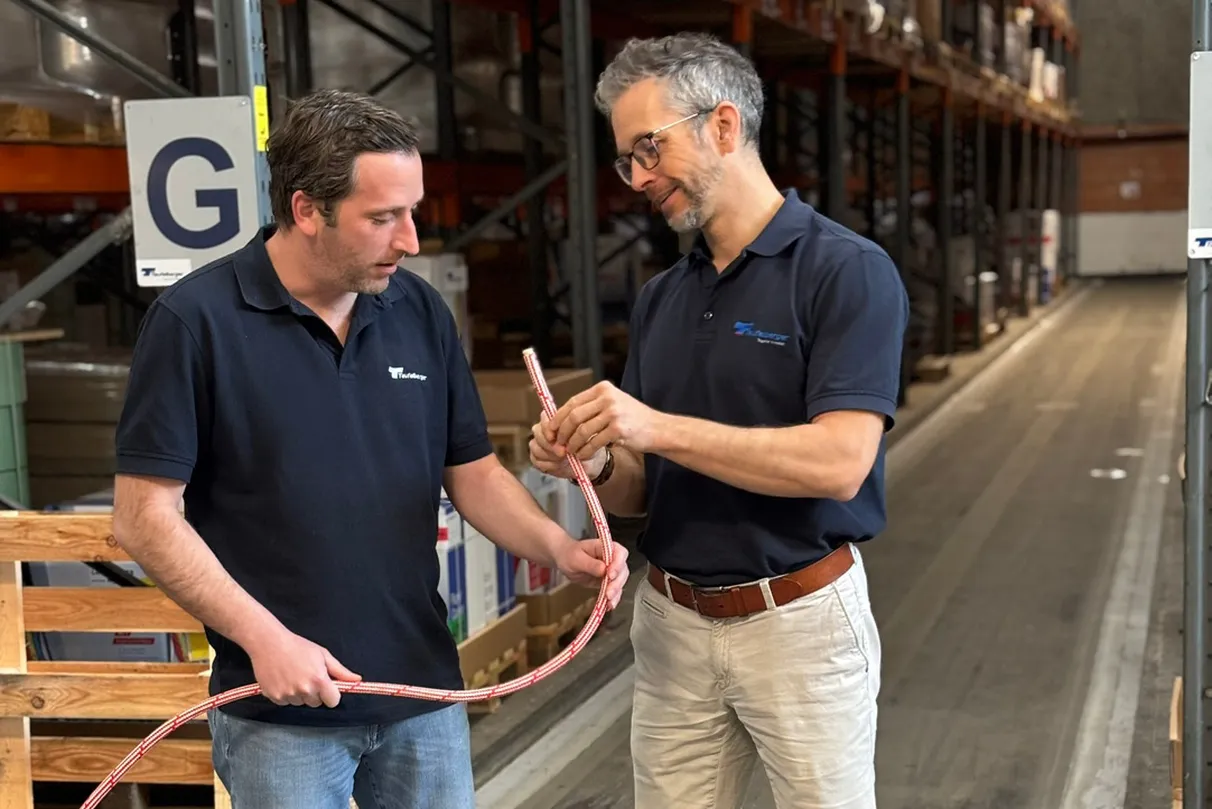 Two individuals in a warehouse setting demonstrating how to handle and tie a rope, with shelves of supplies and a wooden pallet visible in the background.