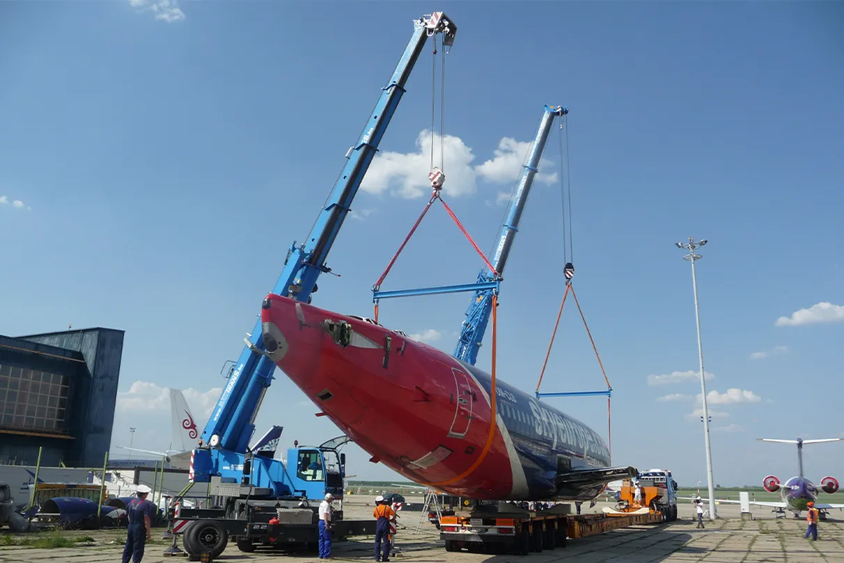Two blue heavy-duty cranes lift the fuselage of a passenger aircraft onto a special transporter at an airport under clear skies