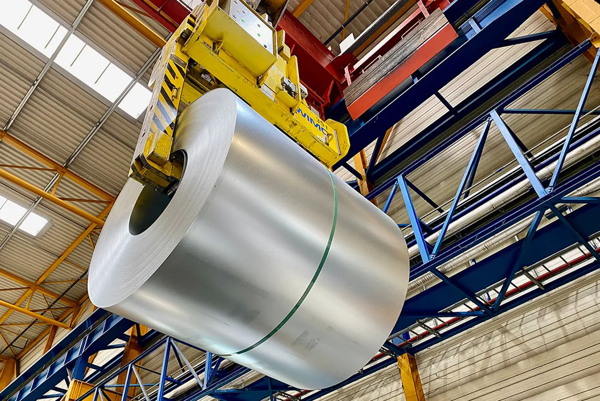 Large metal coil with a shiny surface, secured with green strapping, being lifted by a yellow-black striped industrial crane in a hall with blue and yellow beams.