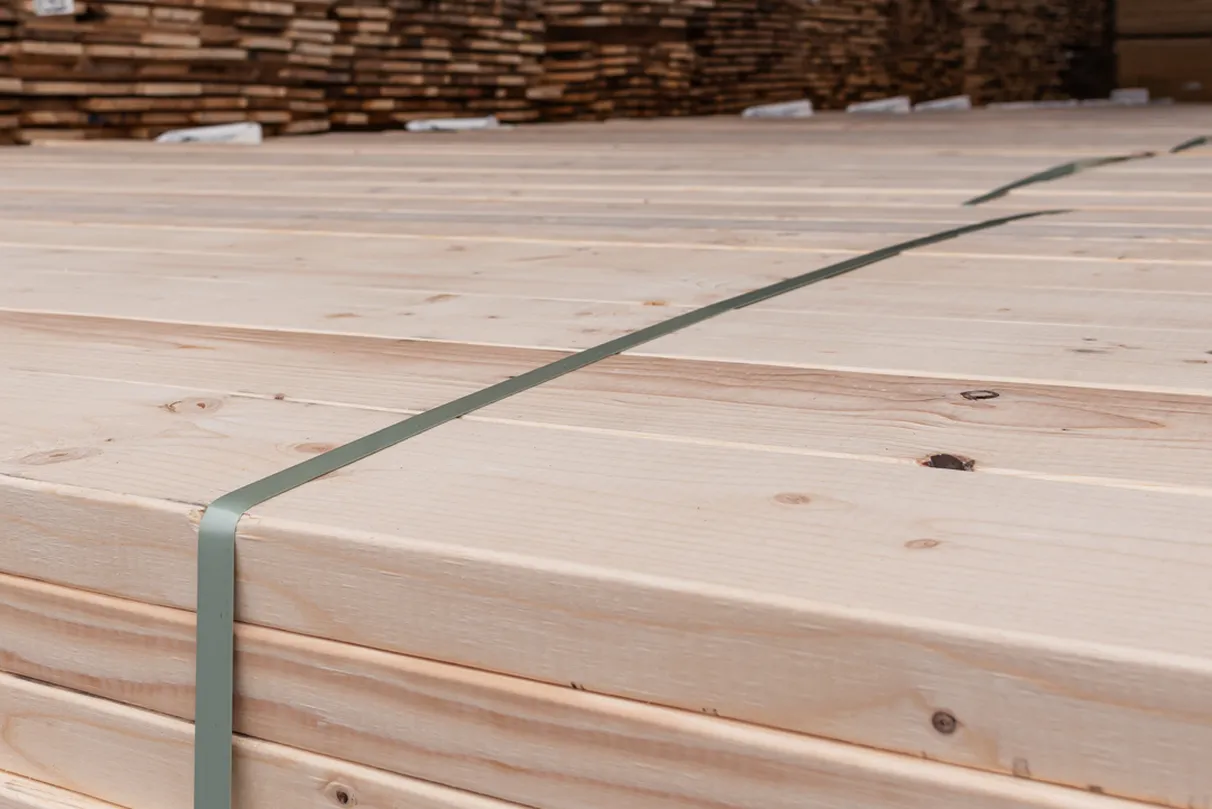 Close-up of stacked light-colored wooden planks with visible grain and knots; bound with a green plastic strap, additional wood stacks in the background.