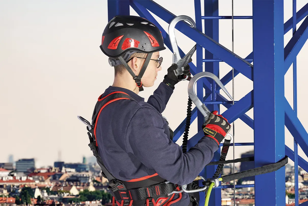 An industrial climber wearing a helmet and safety harness works on a blue metal structure above an urban landscape. The scene highlights professional height work with proper safety measures.