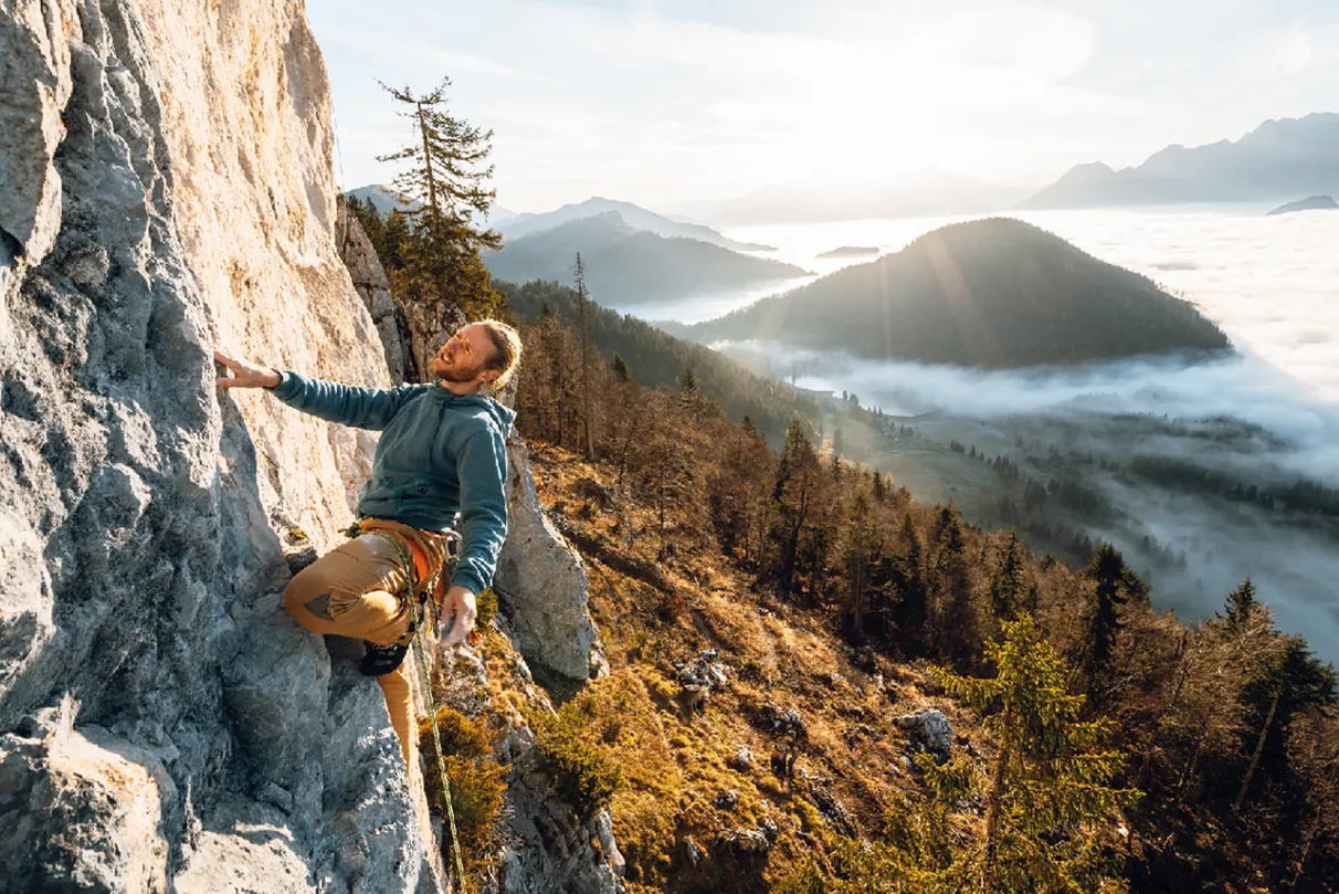 A climber ascends a rocky cliff with a scenic view of mountains, trees, and a misty valley under a bright sun.