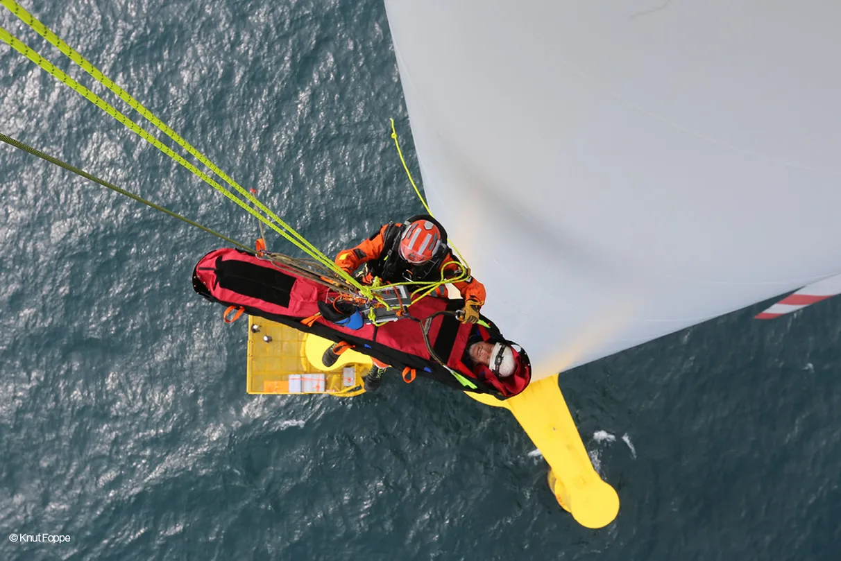 Two rescue workers conduct a high-altitude rescue on a wind turbine above the ocean, securing a person in a red stretcher with yellow ropes. The aerial view emphasizes the complexity and risk of offshore rescue operations.
