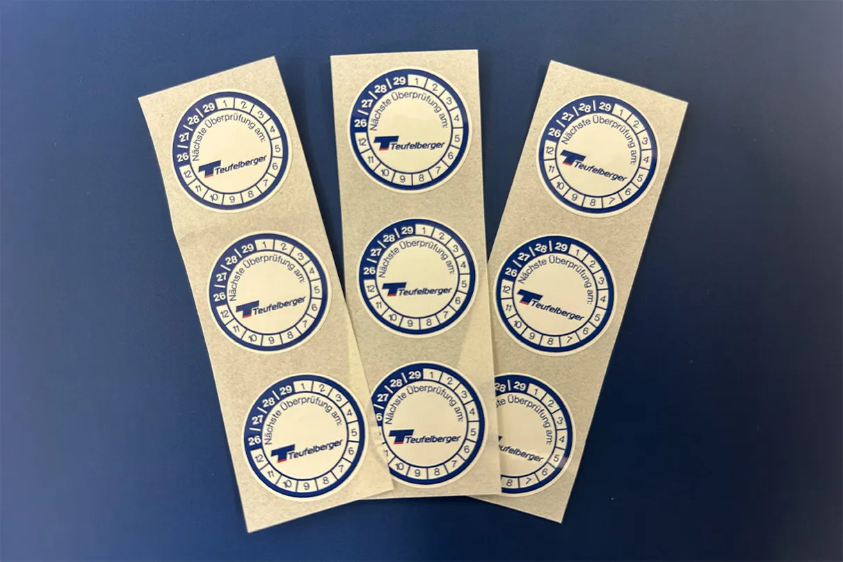 Three sheets of round blue-and-white Teufelberger inspection stickers, each featuring rings for month and year markings used in certification tracking.