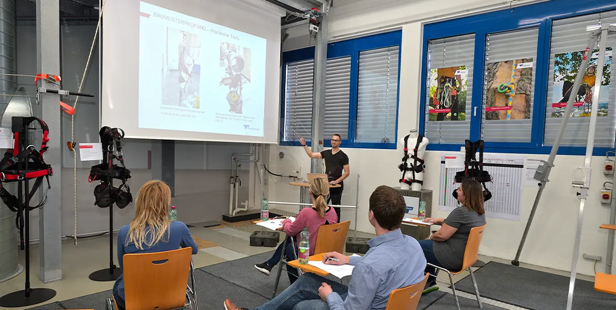 Training room with presenter showing safety harness and rigging slides to seated trainees, demonstrating fiber ropes, wire ropes and strapping quality