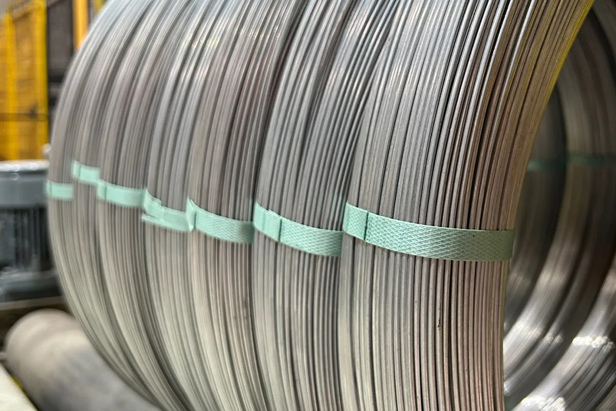 Close-up of large coils of metal wire, bundled with green strapping bands; industrial equipment and structures visible in the background.