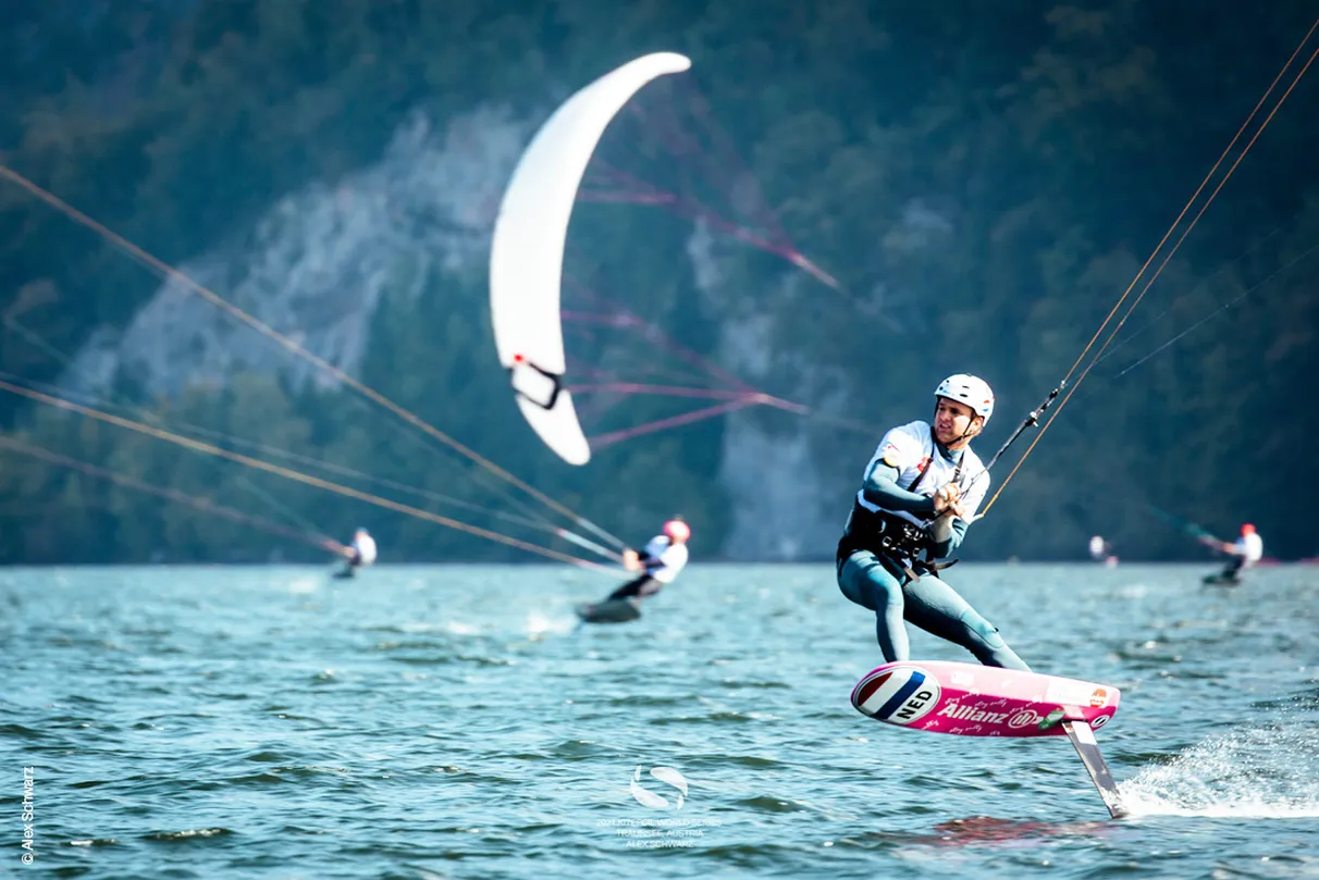 Person kiteboarding on a lake, with others in the background. Mountains and trees are visible in the distance.