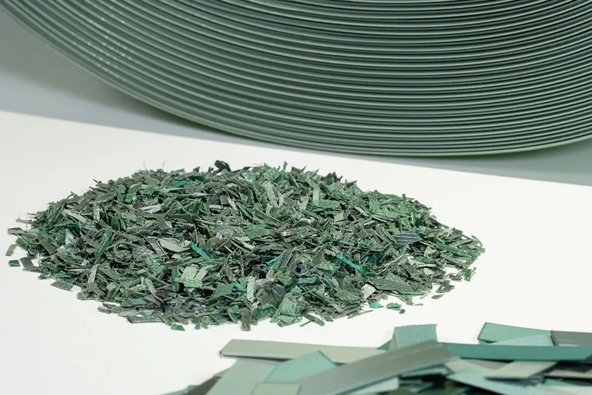 Pile of shredded green material on a white surface, with stacked coil of strapping in the background and unshredded straps in the front.